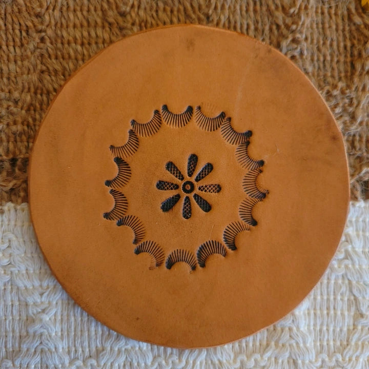 Single leather coaster in medium stain showing a stamped circular starburst border with floral center pattern.