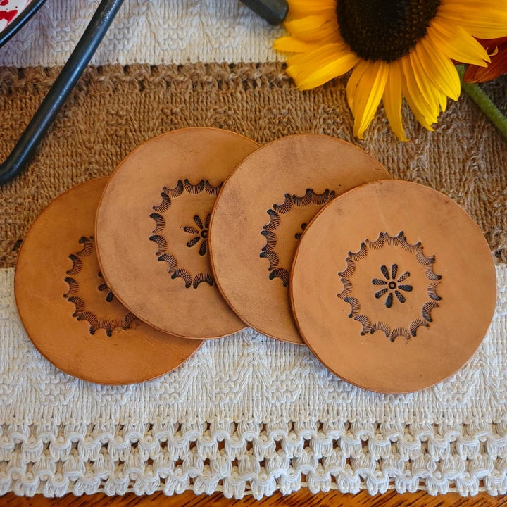 Set of four round leather coasters in medium stain, featuring a stamped starburst circle with floral design in the center.