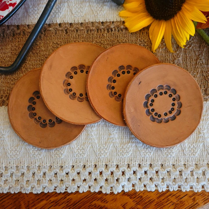 Set of four handmade leather coasters with a circular seashell border design, medium stain, displayed on rustic fabric with a sunflower.