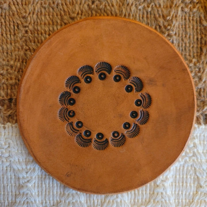 Single handmade leather coaster with a circular seashell border design, medium stain, shown from the front.